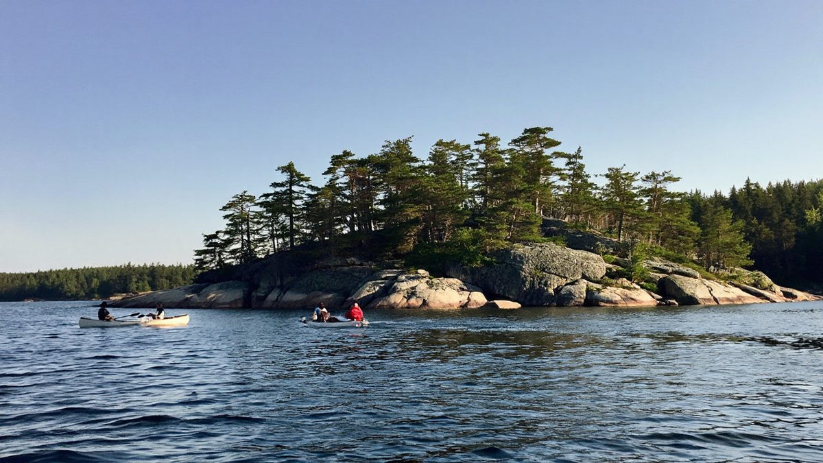 Canoes paddling past a rocky pine-covered island on a deep blue northern lake